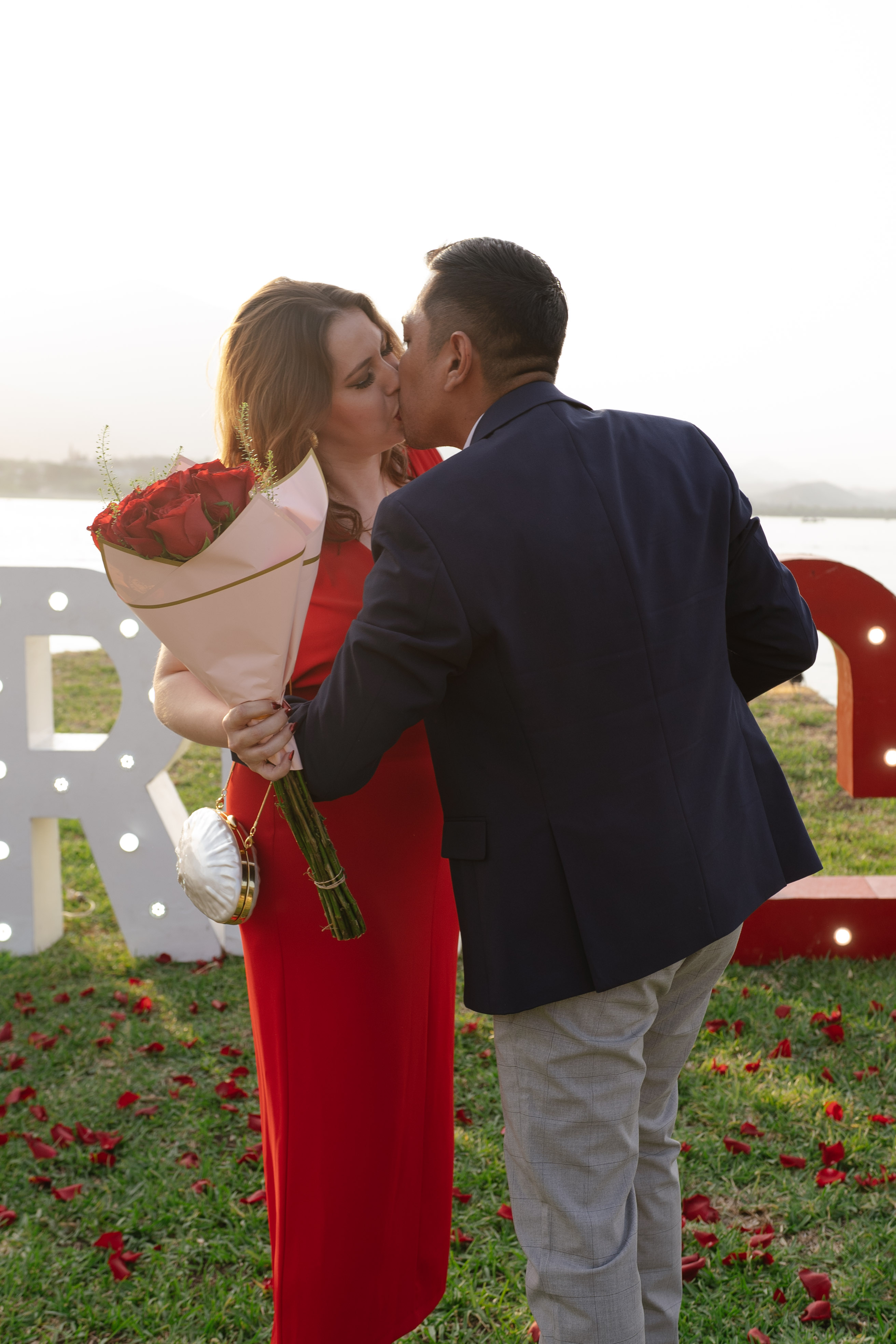 Pareja besándose con vestido rojo y ramo de rosas frente al lago de Monterrey