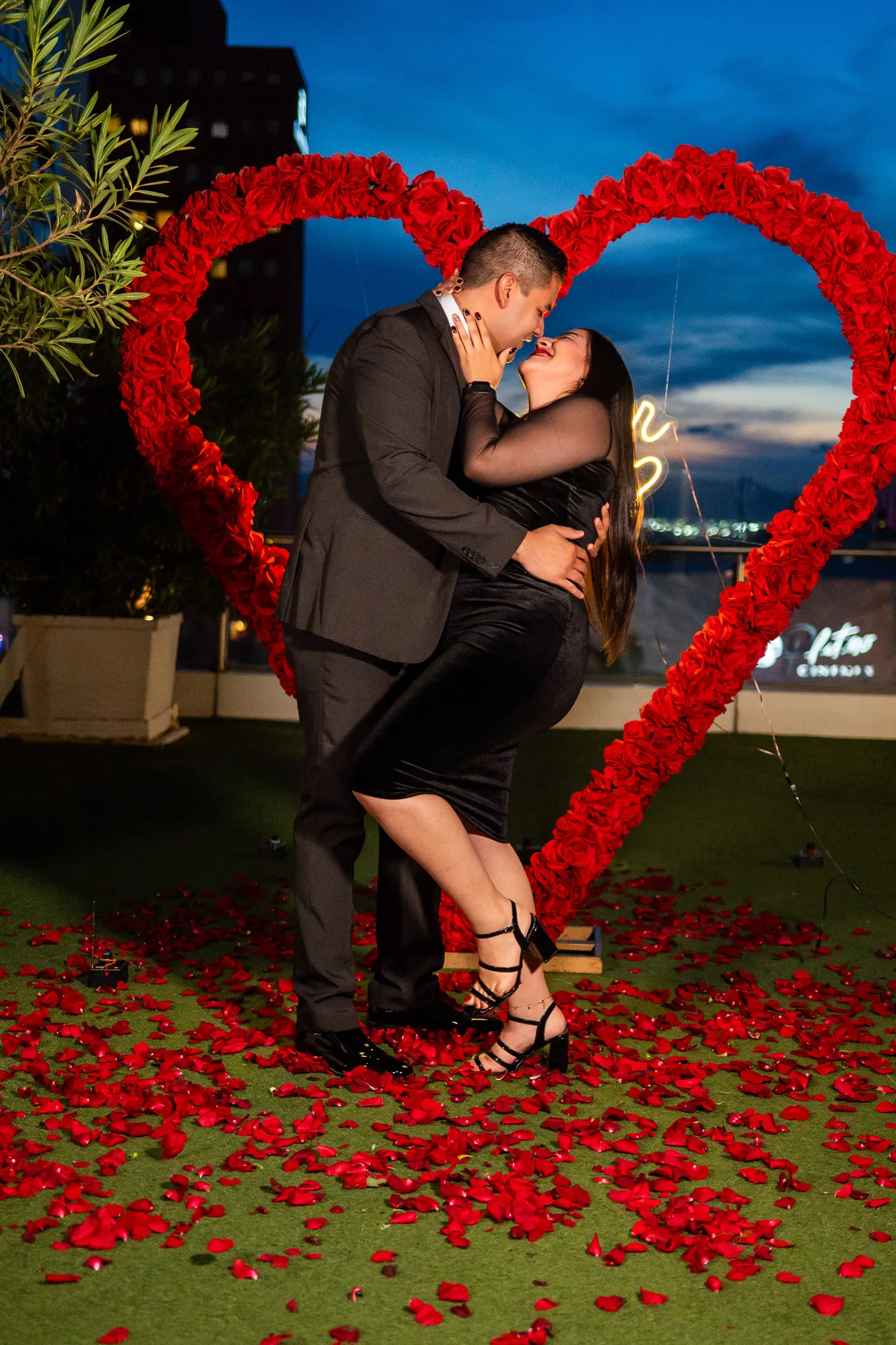 Pareja besándose dentro del arco de rosas rojas de noche, pétalos en el suelo